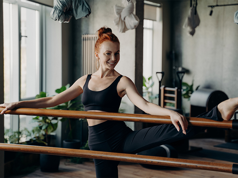 Fitness Trend Barre Workout: Junge rothaarige Frau stretcht mit einem Bein über eine Ballett-Stange und lächelt.
